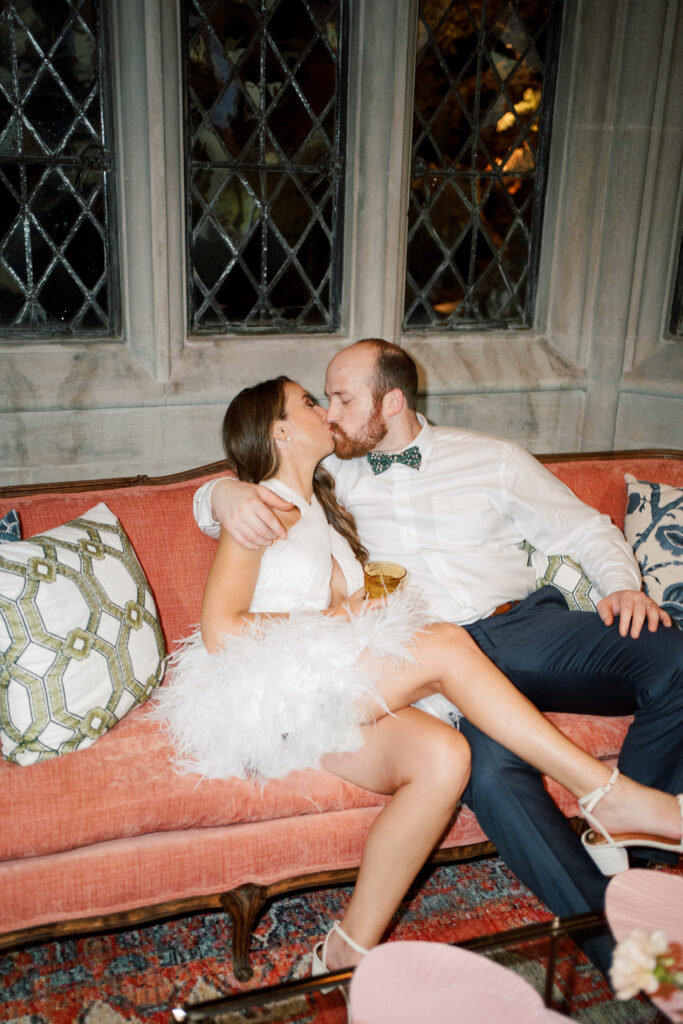 Chic and playful wedding reception moment at The Branch Museum with stylish Richmond couple. Bride and Groom on vintage couch kissing. Bride is in feather mini dress.