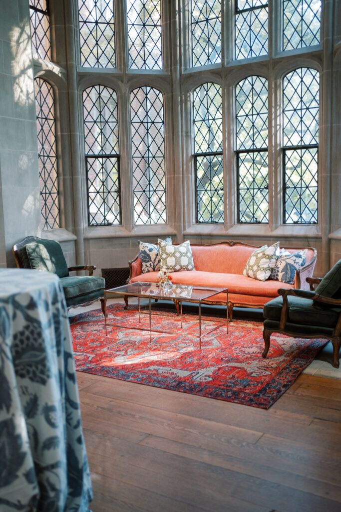 Lounge seating setup inside The Branch Museum featuring a coral velvet sofa, vintage armchairs, and colorful throw pillows beneath leaded glass windows. Branch Museum Wedding, Richmond Va