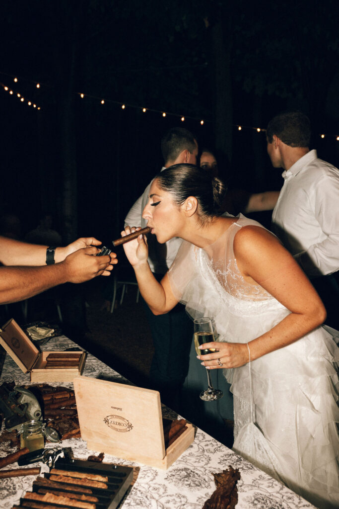 Bride lighting a cigar while holding champagne at an outdoor cigar lounge during her Meridian House wedding reception.