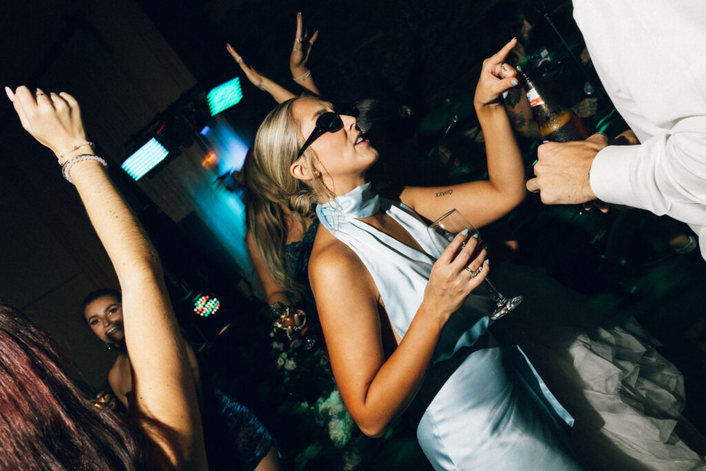 Bridesmaid in blue silk dress wearing sunglasses while dancing with a drink at the Meridian House wedding reception.