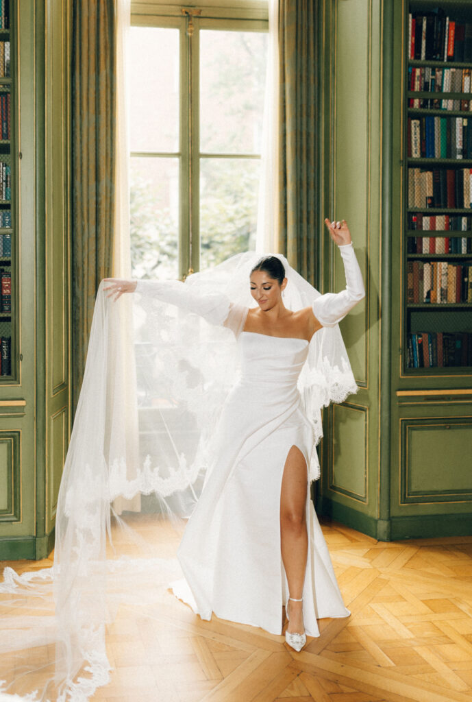Bride in a modern white gown with puff sleeves and a dramatic slit, dancing in the Meridian House library with lace veil draped around her.