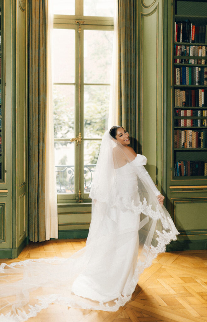 Bride smiling over her shoulder with a sweeping lace veil in the green Meridian House library, framed by French windows.