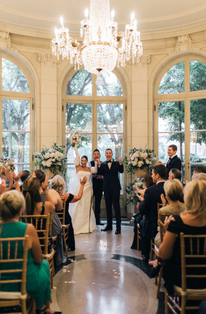 Bride and groom cheer at the altar during their ceremony at Meridian House, surrounded by florals and guests clapping beneath crystal chandeliers. Indoor wedding ceremony at Meridian House with white, light blue, blush, and green florals. Meridian House ceremony rain plan. 