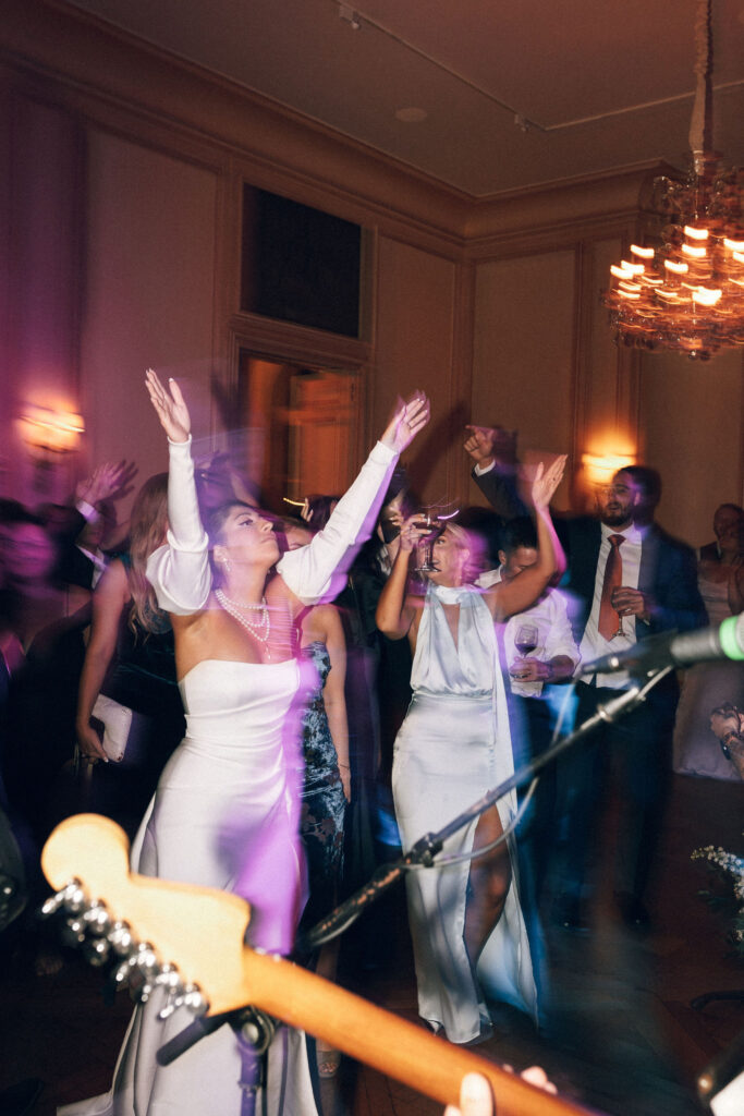 Bride in a modern off-shoulder gown dancing with guests under purple party lighting at Meridian House wedding reception. Dancing to a live band.