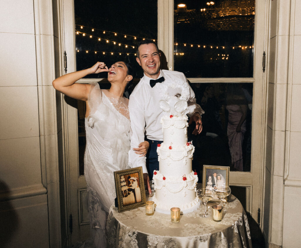 Bride eating a cherry, laughing beside their whimsical cherry-topped wedding cake at Meridian House during a stylish D.C. wedding reception.