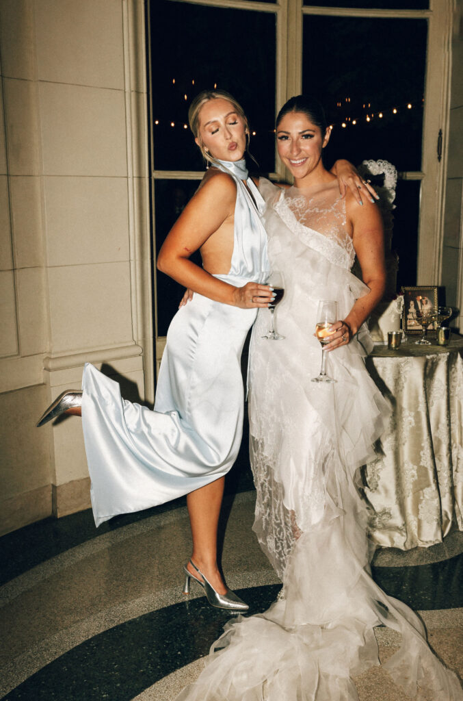 Bride and bridesmaid in blue silk or satin bridesmaid dress pose playfully with champagne flutes during wedding reception at Meridian House.