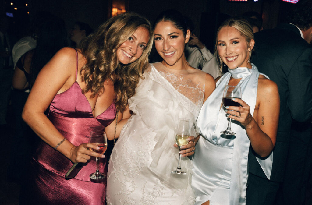Bride with two female guests in jewel-toned evening wear, smiling and holding wine during the Meridian House wedding reception.